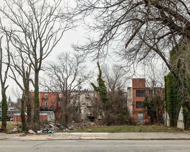 A vacant, overgrown lot sits between brick apartment buildings under a cloudy sky, with bare trees and scattered debris in the foreground.