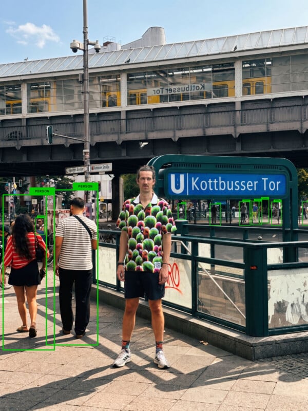A man in a cactus-print shirt stands on a sidewalk in front of a Kottbusser Tor subway entrance, with two people walking away from the camera nearby.