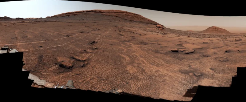 A panoramic view of a rocky, barren Martian landscape with a large, layered mountain in the distance and rover equipment in the foreground.
