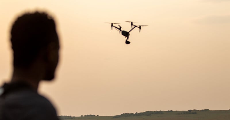 A person in the foreground, seen in silhouette, watches a drone flying in a hazy, golden sky above a distant landscape.