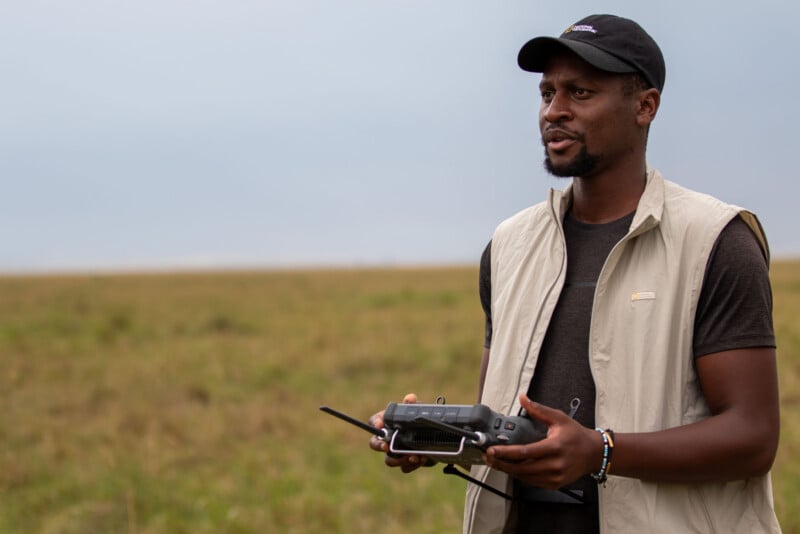 A man wearing a National Geographic cap and a light-colored vest holds a drone remote control while standing in a field.