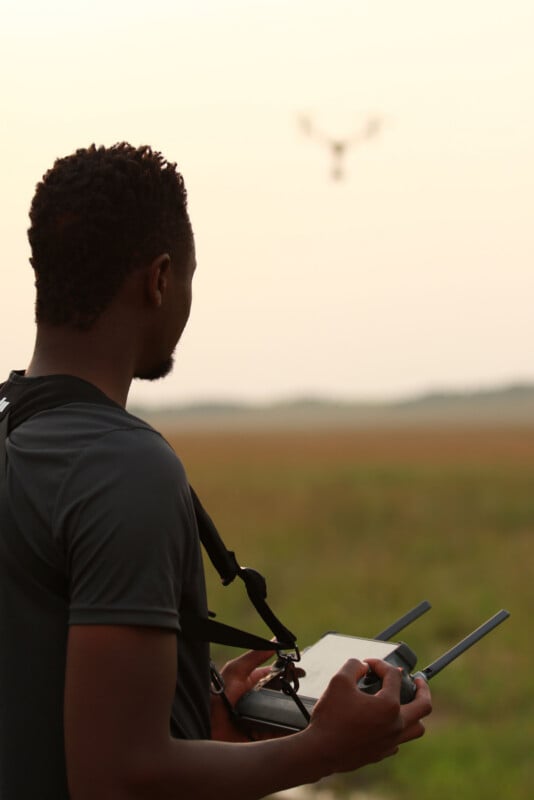 A man seen from behind holds a drone remote control while looking toward a drone flying in the distance over a field.