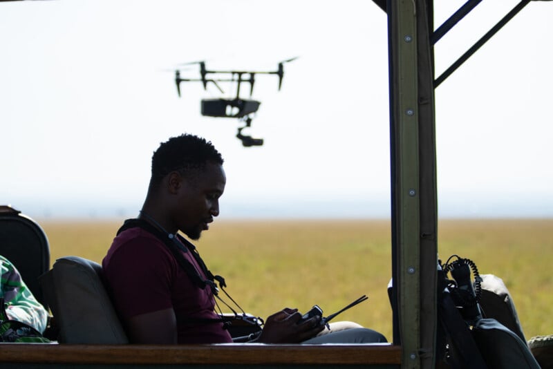 A man in a maroon shirt sits in a vehicle, looking down at a remote control while a drone hovers in the background.