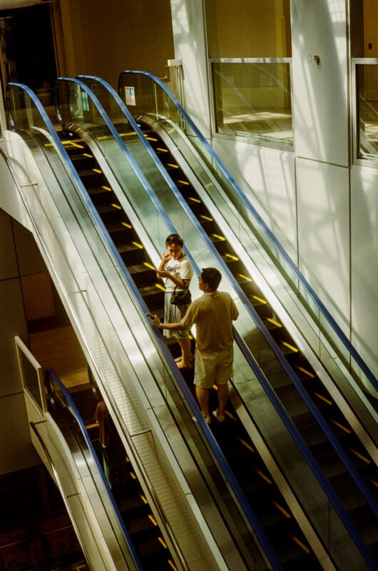 A man and a woman stand on an escalator in a modern building, with another person partially visible on a lower level.
