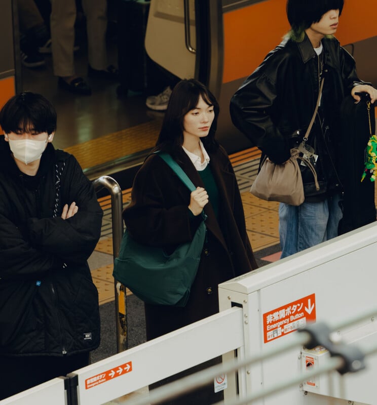 Three people stand on a train platform waiting for a subway train, with a woman in a dark coat and green bag in the center.