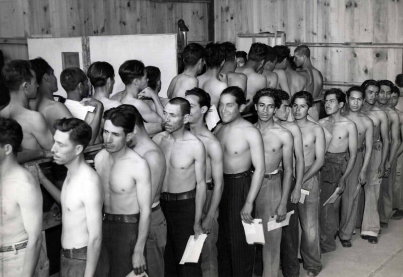 A line of shirtless men standing in a wooden building, many holding small white papers, waiting for a medical or physical examination.