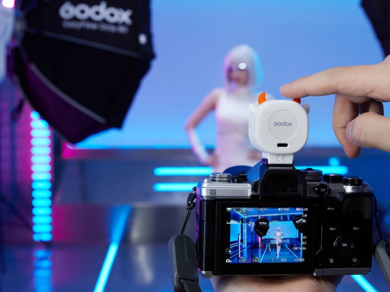 A hand presses a small, white, devil-horned Godox wireless transmitter mounted on top of a digital camera, which displays a live view of a model in a studio.