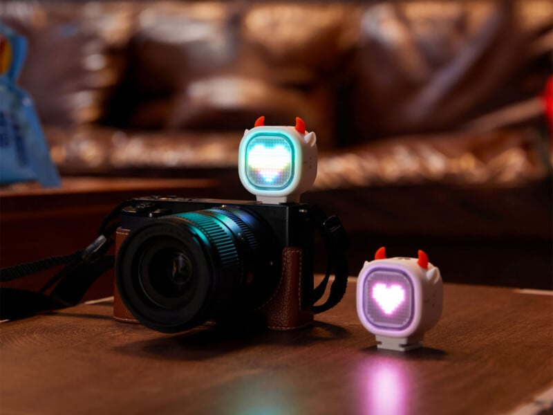 A black mirrorless camera sits on a wooden surface with a small, white, devil-horned LED light attached to its hot shoe, while a matching light sits on the table beside it.