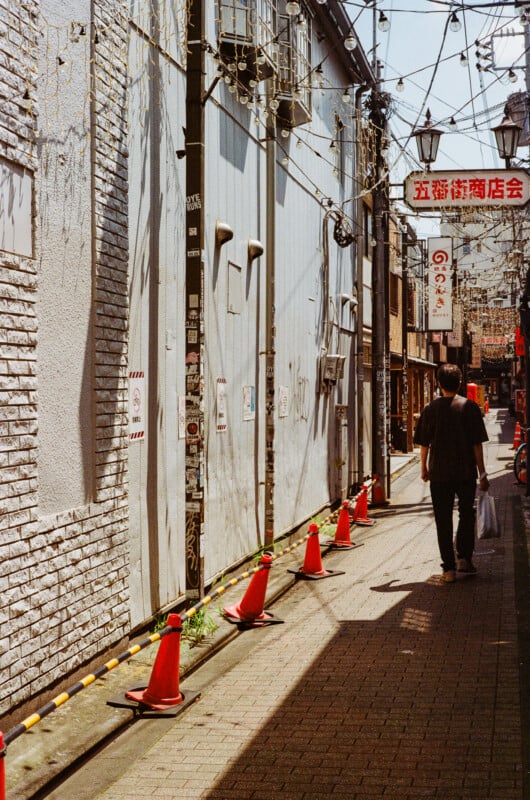 A person walks down a narrow, sunlit alleyway lined with orange traffic cones and a brick wall covered in stickers.