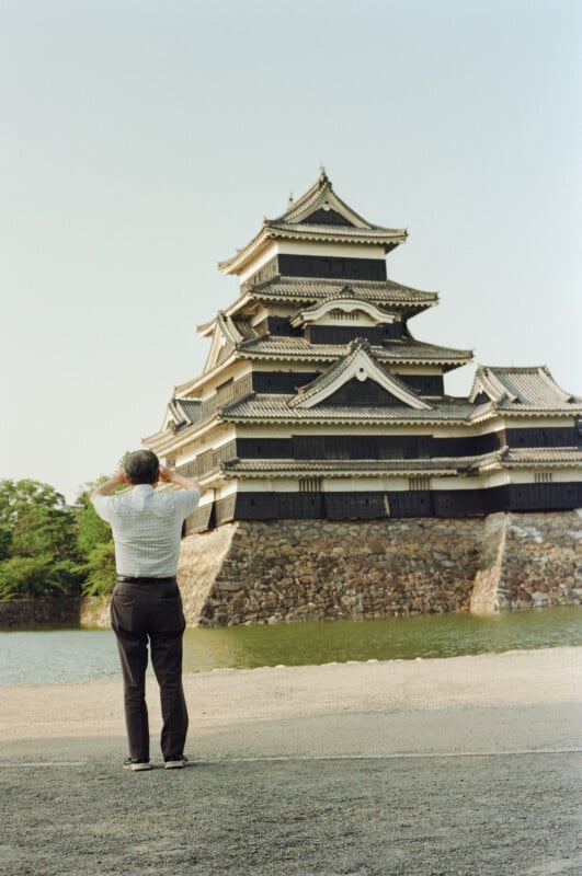 A man in a light-colored short-sleeved shirt and dark trousers stands with his hands raised to his head, looking at a large, multi-storied black castle across a moat.