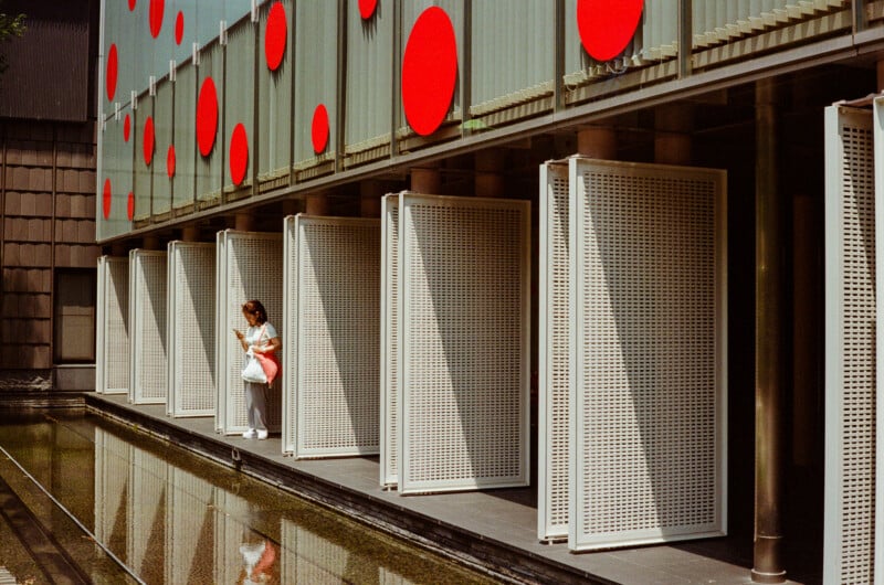 A woman stands on a walkway beside a reflecting pool, looking at her phone in front of a building facade decorated with large red circles and white perforated panels.