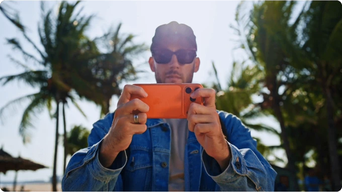 A man wearing a baseball cap and sunglasses holds an orange smartphone horizontally in front of his face outdoors with palm trees in the background.