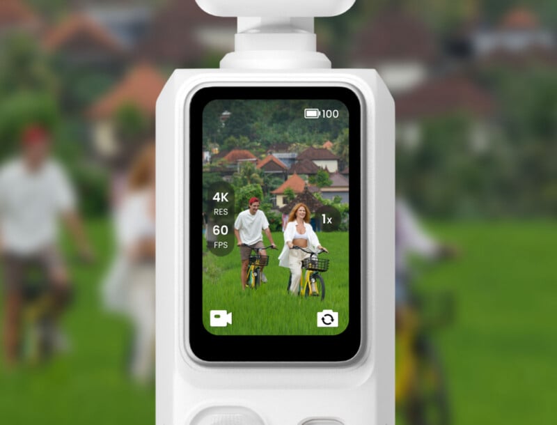 A close-up of a white handheld camera with a vertical touchscreen display showing a live view of a man and woman riding bicycles through a green field.