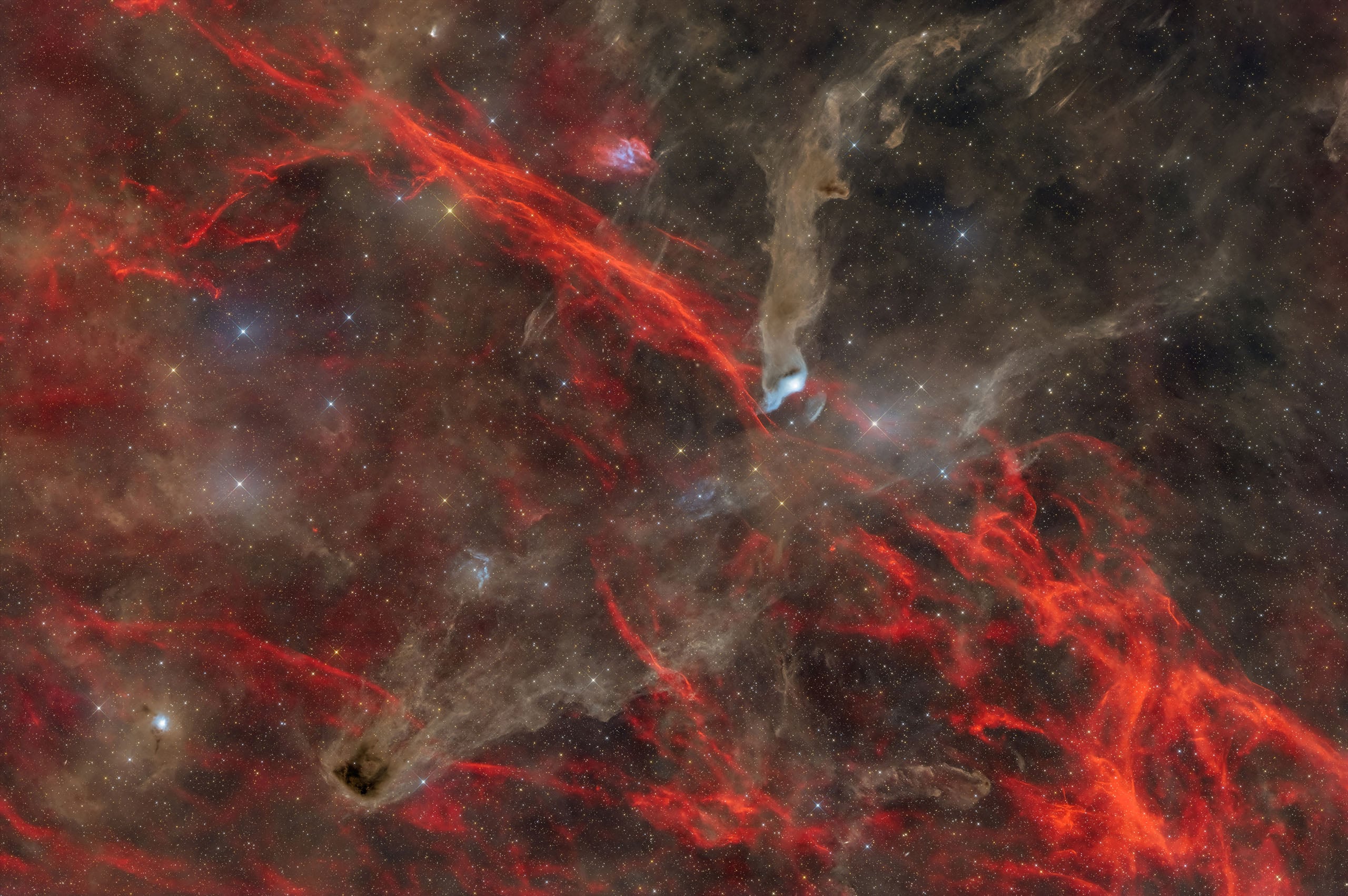 A vast nebula featuring intricate, glowing red filaments of ionized gas set against dark, swirling clouds of cosmic dust.