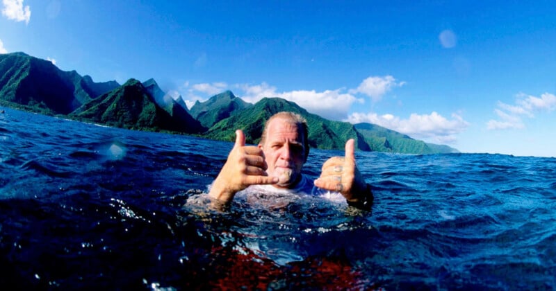 A man in the ocean makes a shaka gesture with both hands while mountains rise in the background under a clear blue sky.