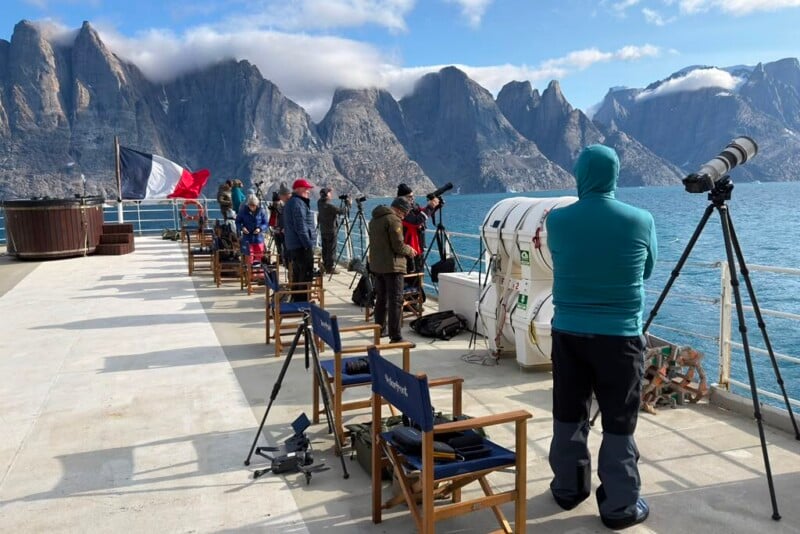 A group of people on the deck of a ship use cameras and spotting scopes on tripods to view a mountainous coastline under a blue sky.