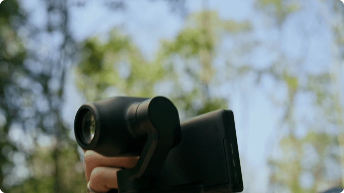A hand holds a black gimbal camera against a blurred background of trees and a bright sky.