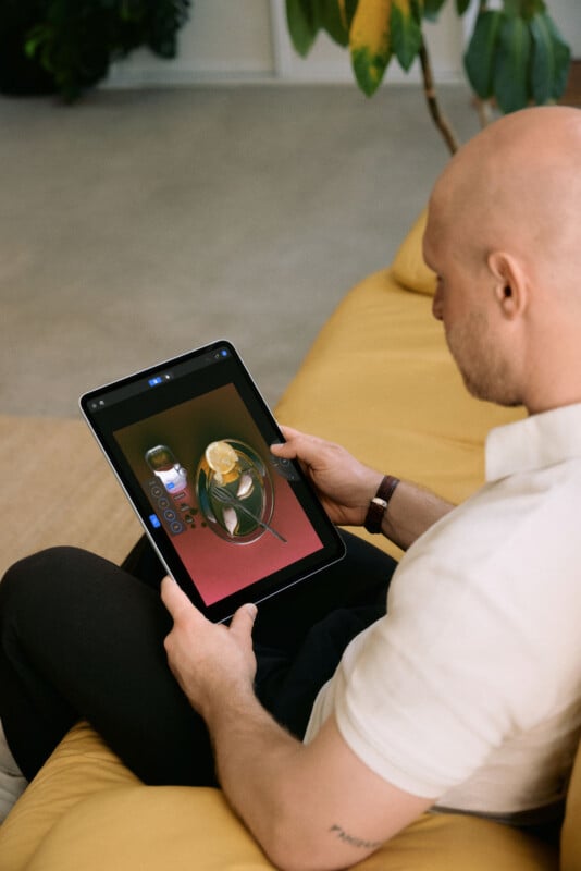 A bald man in a white shirt sits on a yellow sofa, holding a tablet displaying an image editing interface with a bowl of food.
