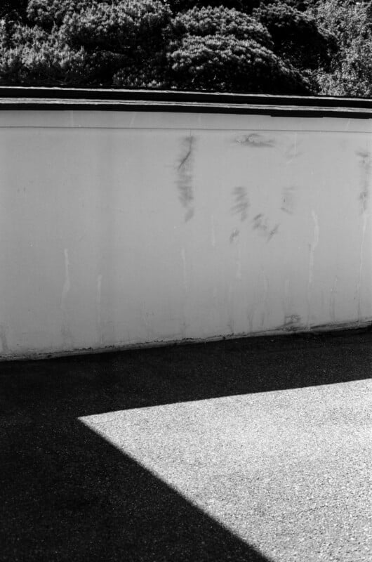 A high-contrast black and white shot shows a plain, weathered wall topped with dark foliage, with a sharp triangular shadow cast across the ground in the foreground.