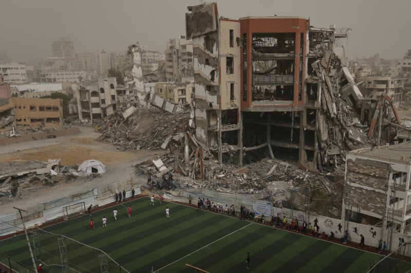 A soccer match is played on a green turf field in the foreground, set against a backdrop of heavily damaged and collapsed buildings.