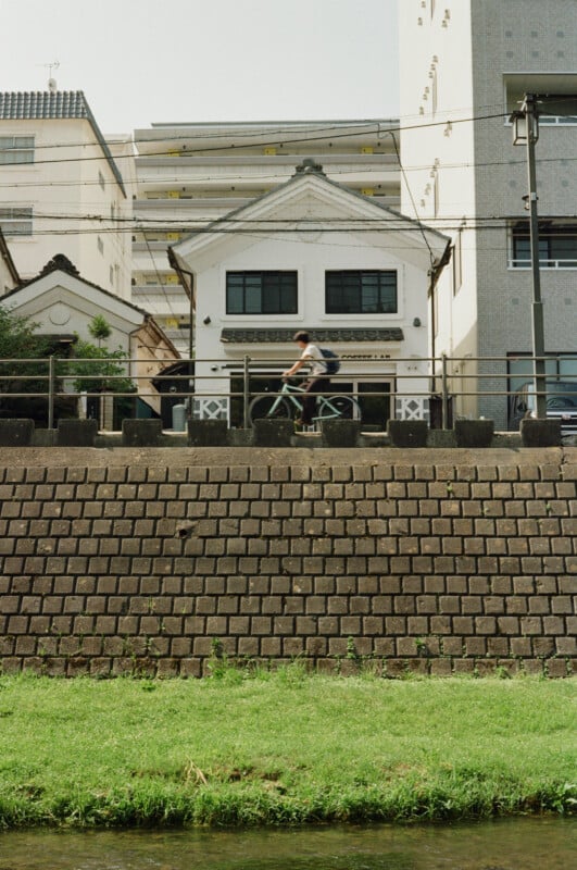A person rides a bicycle along a path in front of a white building with a gabled roof, situated above a stone retaining wall.