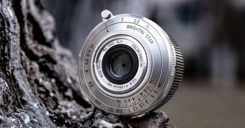 A silver Brightin Star 28mm f/2.8 lens rests on a textured tree bark surface, showing its aperture blades and focus markings.