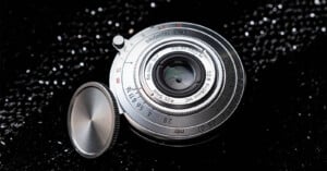 A silver Brightin Star 28mm f/2.8 lens with a detached circular metal lens cap, resting on a black, glittery surface.