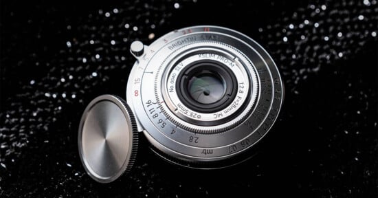 A silver Brightin Star 28mm f/2.8 lens with a detached circular metal lens cap, resting on a black, glittery surface.