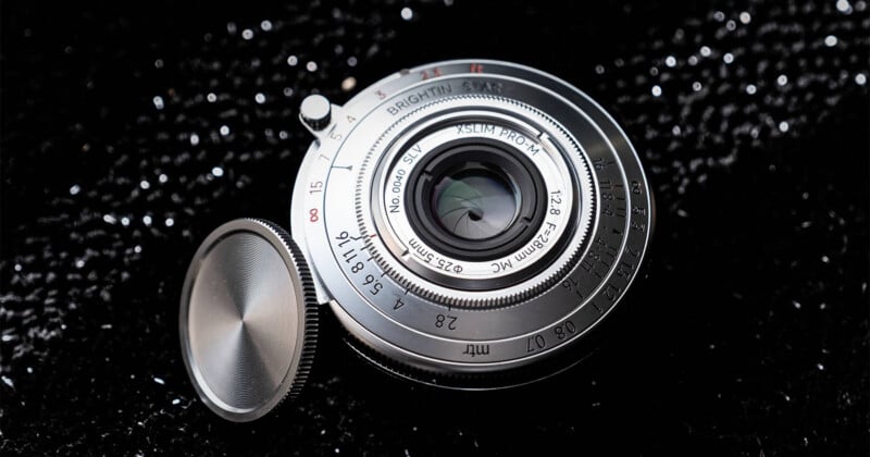 A silver Brightin Star 28mm f/2.8 lens with a detached circular metal lens cap, resting on a black, glittery surface.