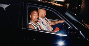 Tupac Shakur sits in the passenger seat of a car next to Suge Knight, who is driving.