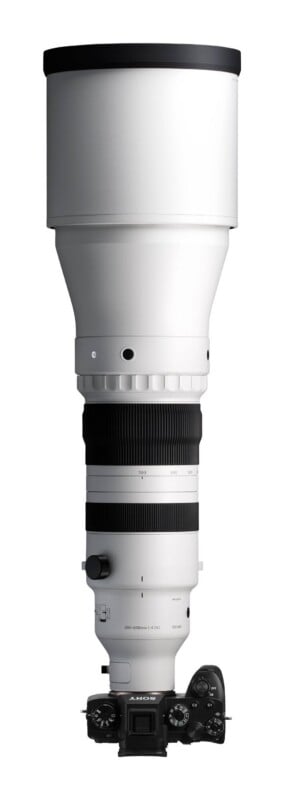 A white telephoto zoom lens attached to a black Sony Alpha mirrorless camera body, shown vertically against a white background.
