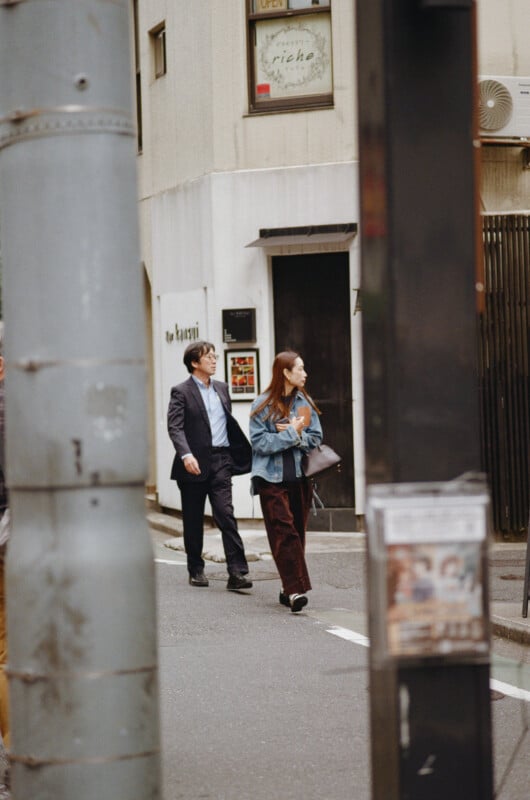 A man in a suit and a woman in a denim jacket and corduroy pants walk along a city street past a building with a sign reading "riche".