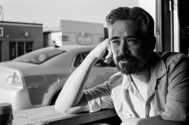 A man with a beard and graying hair rests his chin on his hand while looking out a window, with a car visible in the background.