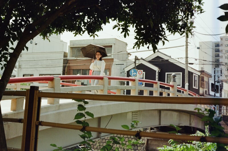 A woman holding a black umbrella stands on a white bridge with red railings, with buildings and utility poles in the background.