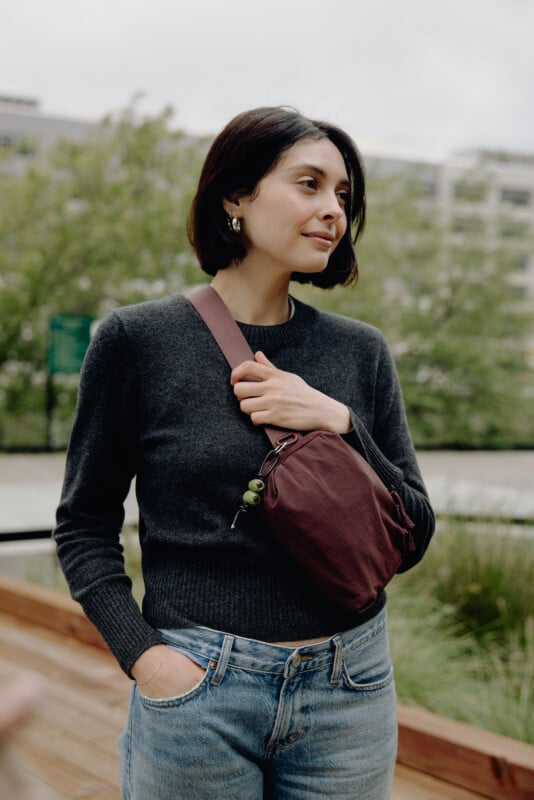 A woman with short dark hair wears a dark gray sweater, blue jeans, and a maroon crossbody bag while standing outdoors.