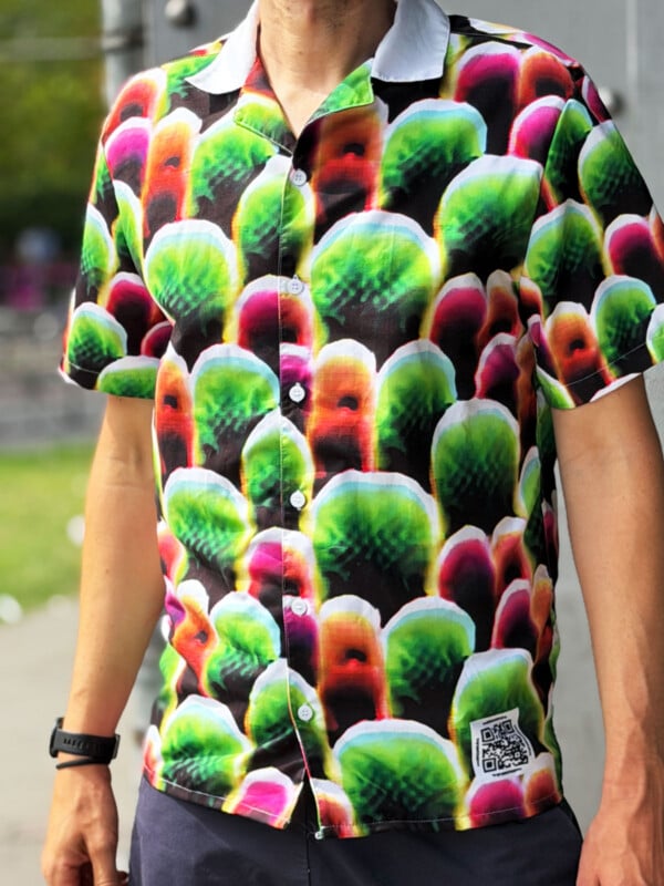 A person wears a short-sleeved button-up shirt with a repeating pattern of green, red, and white rounded shapes on a black background, featuring a white collar and a small QR code patch on the lower front.