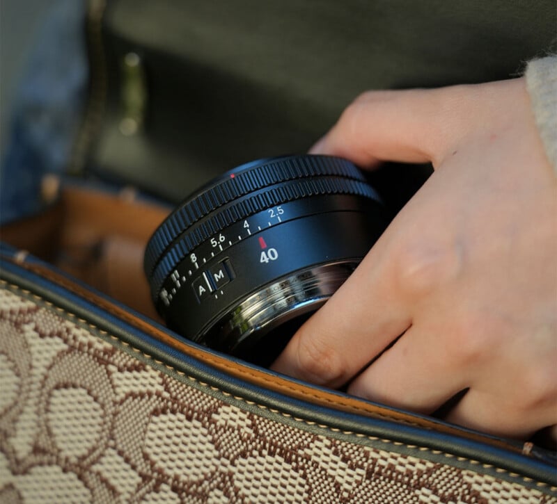 A hand places a black camera lens marked with "40" and an "A/M" switch into a patterned handbag.