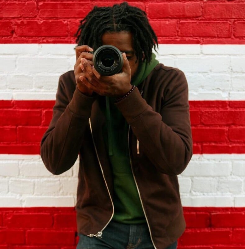 A person with dreadlocks wearing a brown zip-up hoodie over a green shirt holds a camera up to their face in front of a red and white striped brick wall.