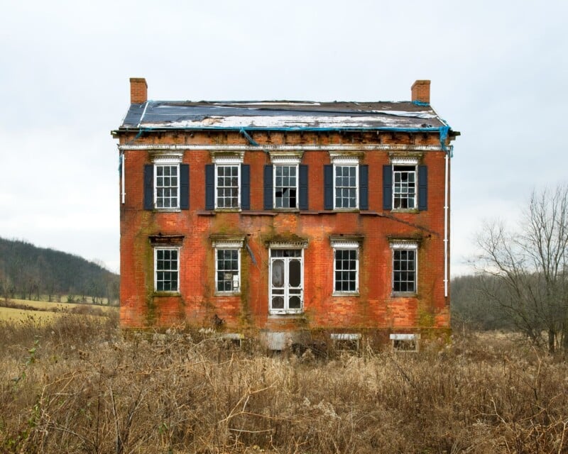 A two-story, weathered red brick house stands in a field of dry, brown grass under an overcast sky, with blue tarps covering its damaged roof.