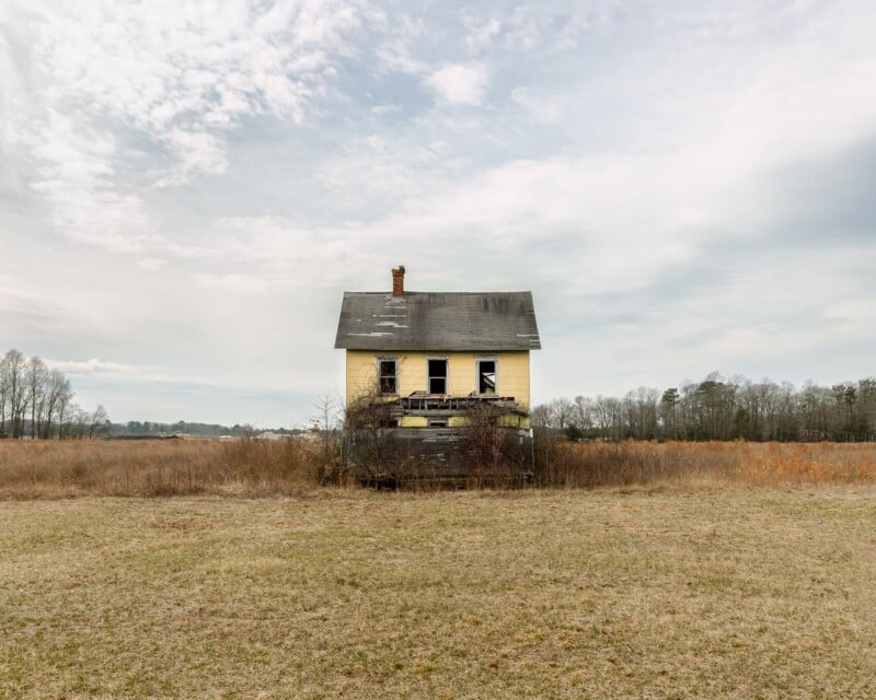 A weathered, yellow two-story house with missing windows and a collapsed porch stands alone in the middle of a vast, dry field under a cloudy sky.