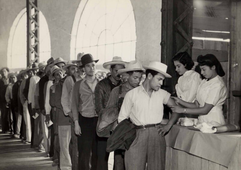 A line of men wearing hats and work clothes wait to receive vaccinations from two nurses at a table.
