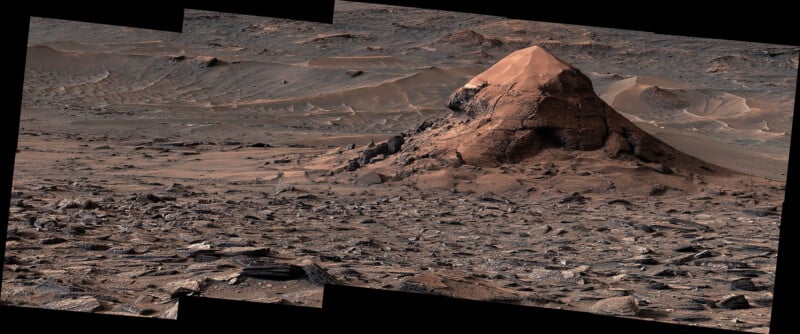 A rocky, barren Martian landscape features a prominent, cone-shaped hill covered in reddish dust surrounded by scattered debris.
