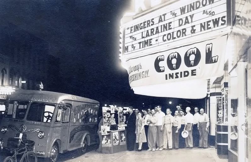 A line of people stands on a sidewalk outside a movie theater at night, with a "Snow Builder" truck parked nearby and a marquee advertising the film "Fingers at the Window.