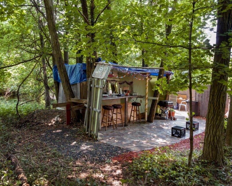 A makeshift outdoor bar with three stools sits under a blue tarp roof in a wooded area, featuring a grill, a fan, and a tiled patio floor.