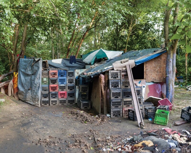 A makeshift shelter constructed from tarps, plywood, and stacked plastic milk crates sits in a wooded area.