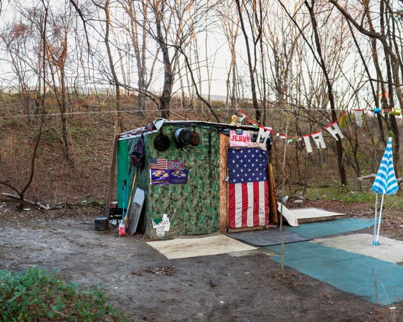 A makeshift shelter constructed from camouflage tarps and wood sits in a wooded area, decorated with a large American flag, Baltimore Ravens logos, and a banner reading "JESUS.