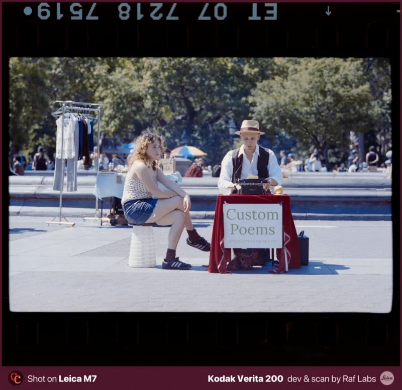A woman sits on a stool next to a man at a table with a typewriter and a sign that reads "Custom Poems" in a public park.