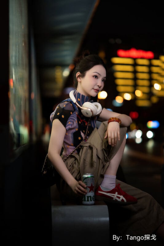 A young woman with her hair in a bun sits on a bench at night, wearing a patterned top, khaki pants, and red sneakers while holding a can of Heineken.