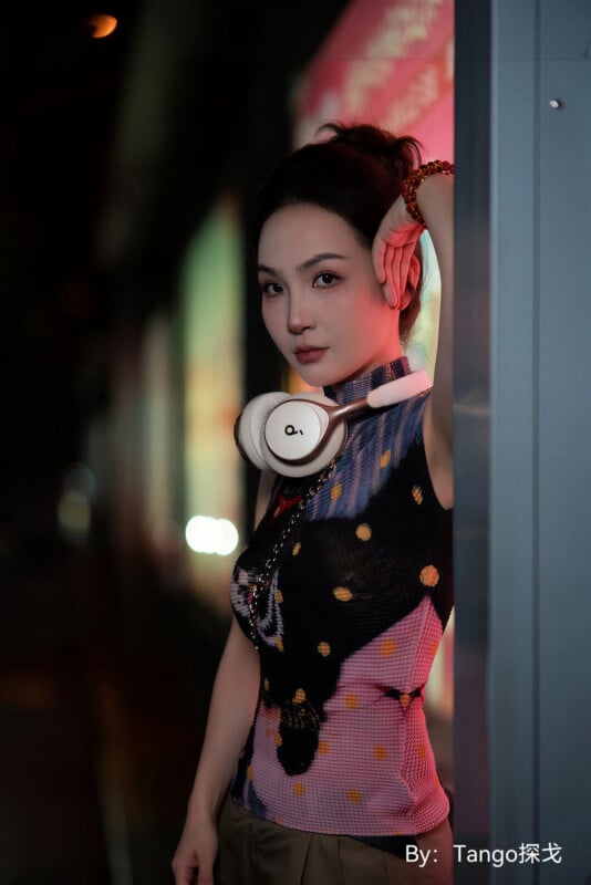 A woman with her hair in a bun wears white headphones around her neck and a patterned, sleeveless top while leaning against a wall at night.