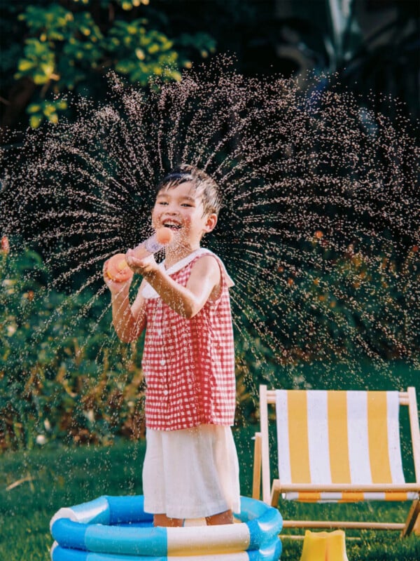A young child in a red and white checkered top stands in a small inflatable pool, laughing while holding a water toy that sprays water in a wide arc.